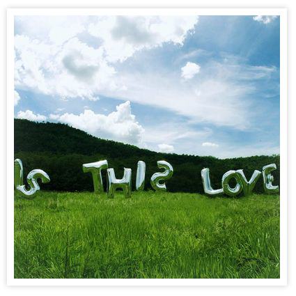 Zielona ��ka, las, b��kitne niebo z chmurami i srebrne balony tworz�ce napis Is This Love